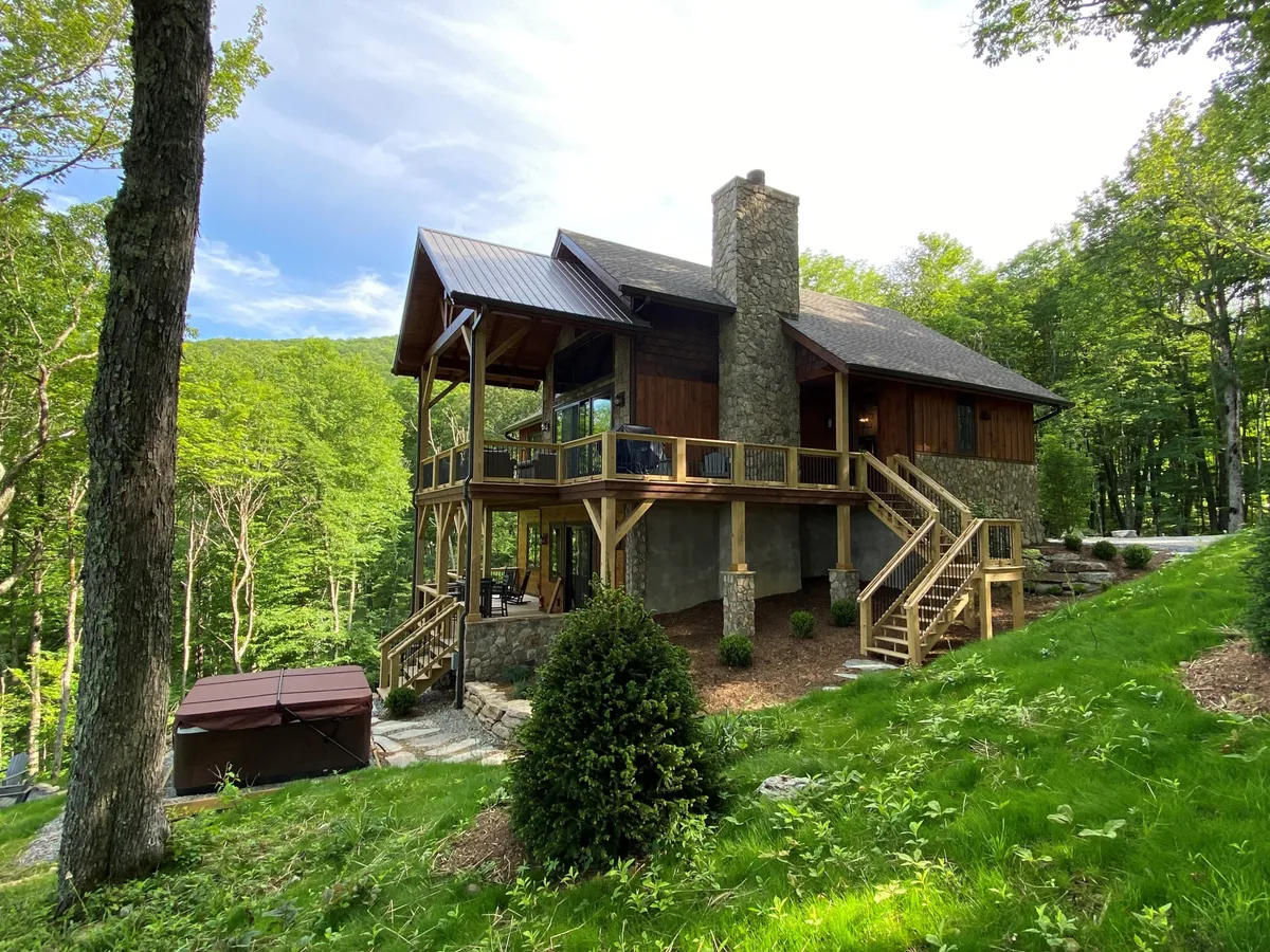 A wooden deck wraps around the home, featuring a hot tub and stone chimney, with trees and greenery all around. The scene suggests a calm spot for relaxing tired muscles after outdoor activities and encourages evening gatherings under the stars.