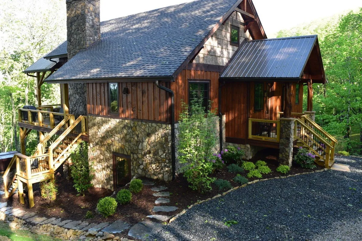 A cozy stone chimney and wooden deck are surrounded by lush greenery, with a stone pathway leading into the landscape. The deck offers ample space for relaxing, outdoor dining, or soaking in the views. The vibrant foliage and inviting setup establish a strong connection to nature and the outdoors.