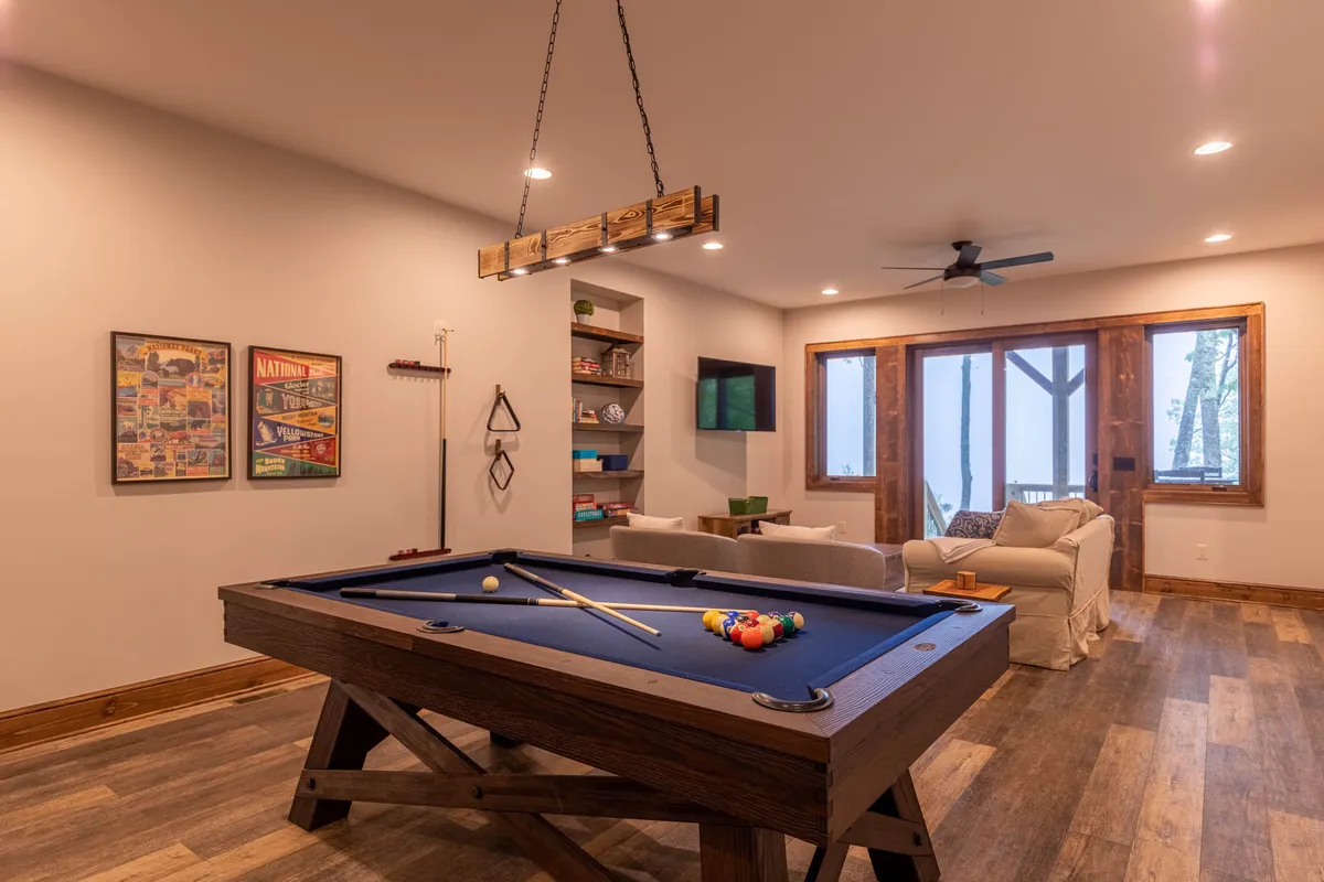 A casual and playful interior with a pool table at the center, a comfortable sofa for lounging, large windows letting in ample natural light, and well-stocked shelves with books. This space is ideal for game nights, reading, or simply unwinding with friends and family.