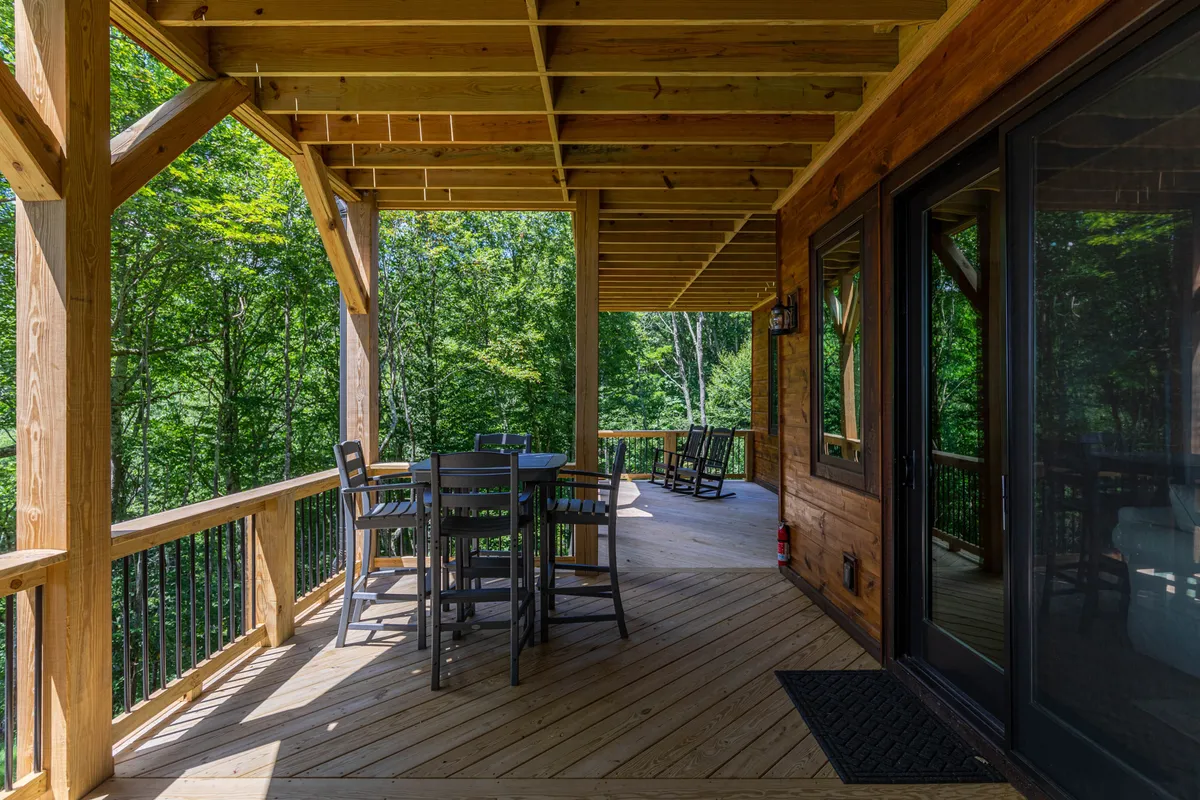 A spacious wooden deck features an outdoor dining table, comfortable rocking chairs, and is shaded by mature trees, creating a tranquil environment ideal for relaxing with pets and enjoying the outdoors.