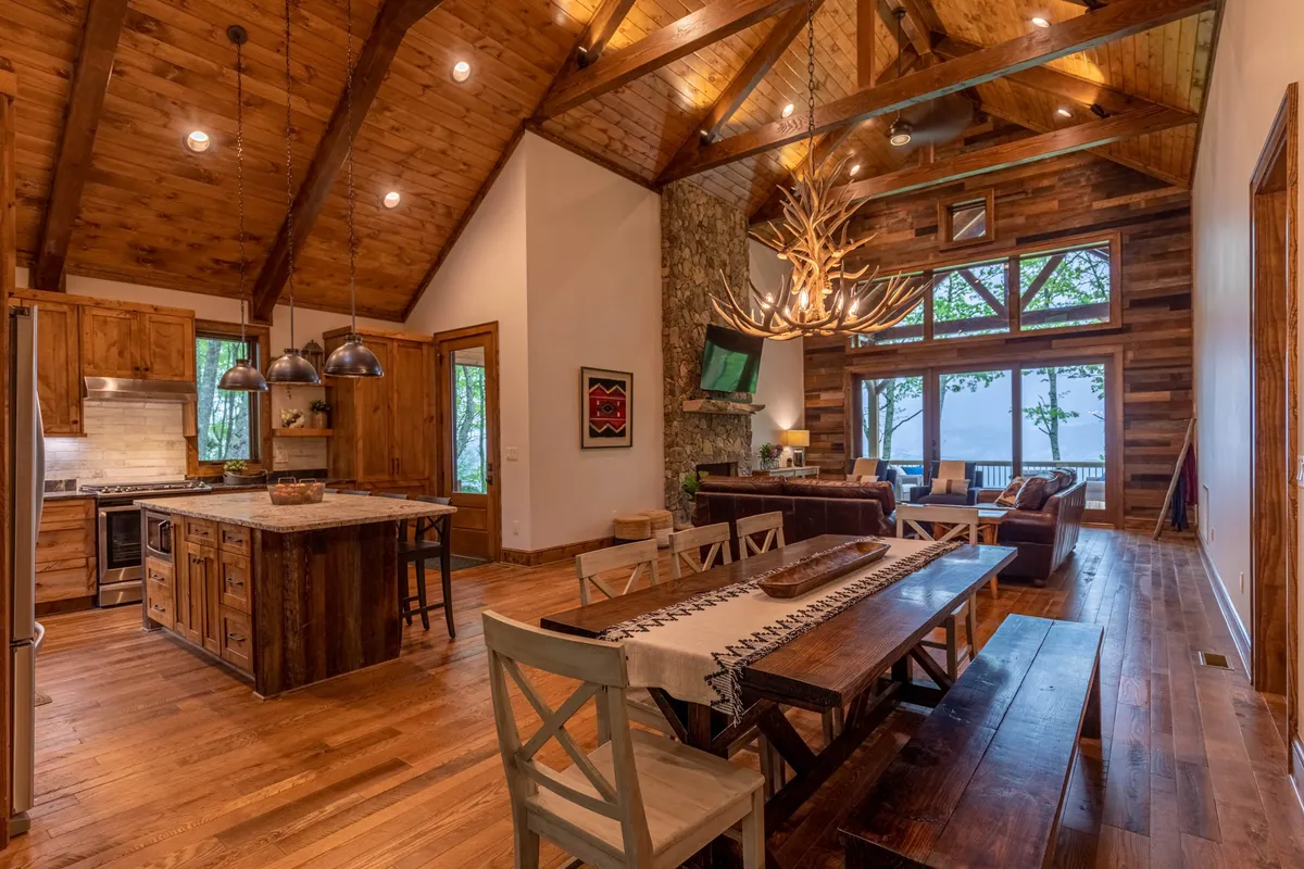 A large rustic dining area with an expansive table set for twelve, illuminated by an antler chandelier. The dining space is adjacent to the kitchen and great room, with large windows framing scenic outdoor views and letting in abundant natural light—perfect for festive gatherings, game nights, or morning coffee with a mountain backdrop.