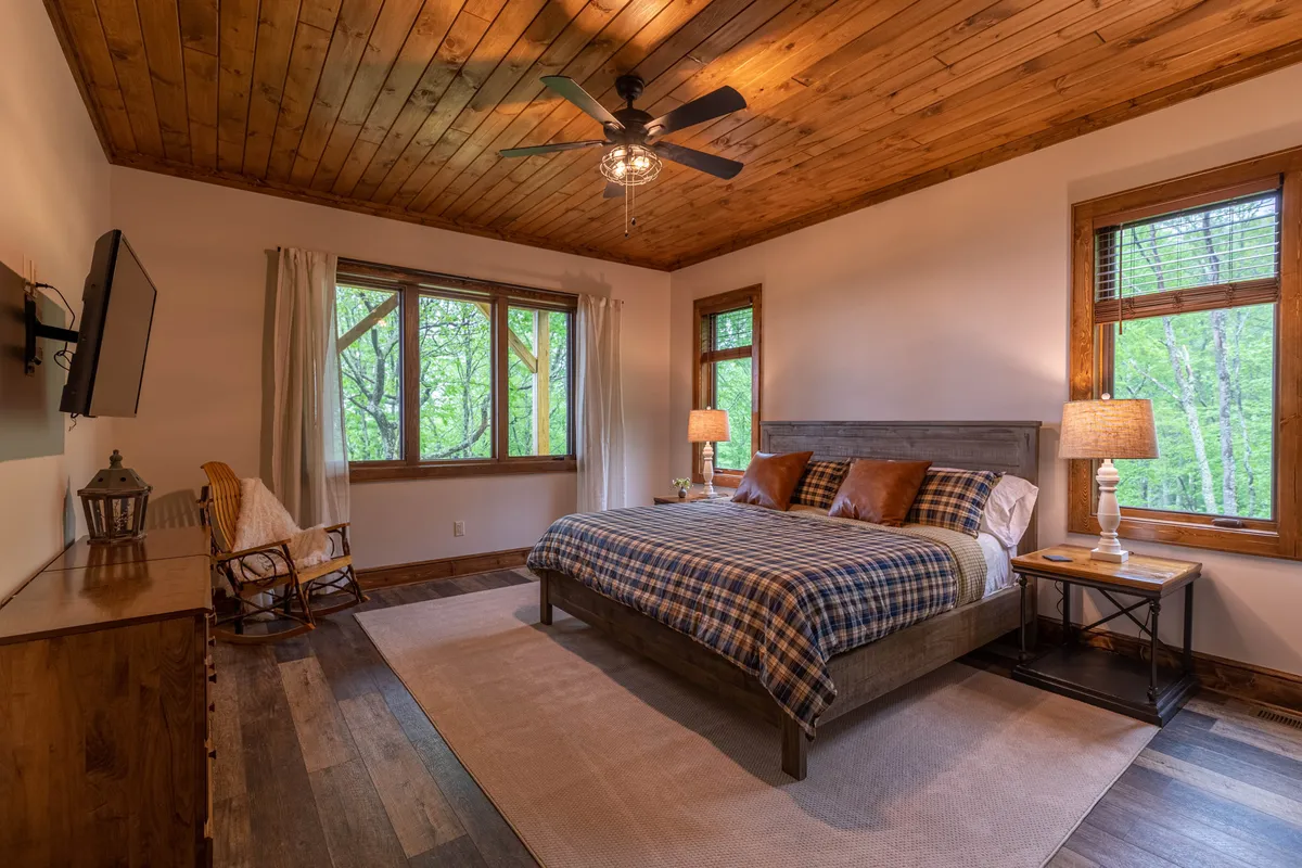 A king bedroom with a plaid bedspread, a wooden ceiling, and large windows. Nightstands with lamps flank the bed, and a cozy rocking chair is positioned nearby, offering a tranquil spot to relax and take in the views. The windows allow natural light to flood the room, highlighting the restful atmosphere.