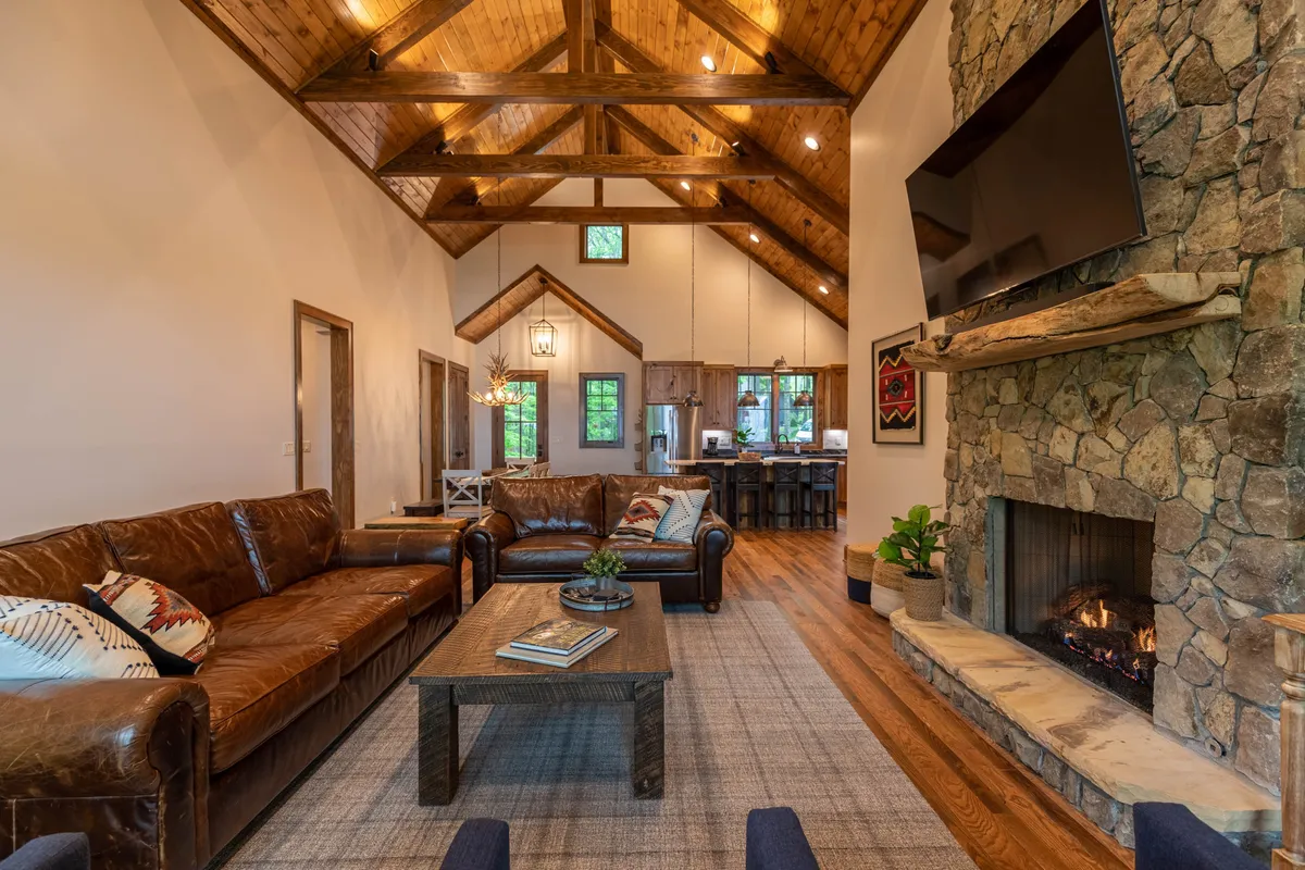 A warm, inviting great room with a soaring vaulted ceiling, exposed wooden beams, and a dramatic stone fireplace as its centerpiece. Plush leather sofas are arranged around the hearth, with ample natural light streaming in through expansive windows. The space feels open yet cozy, perfect for gatherings and relaxation.