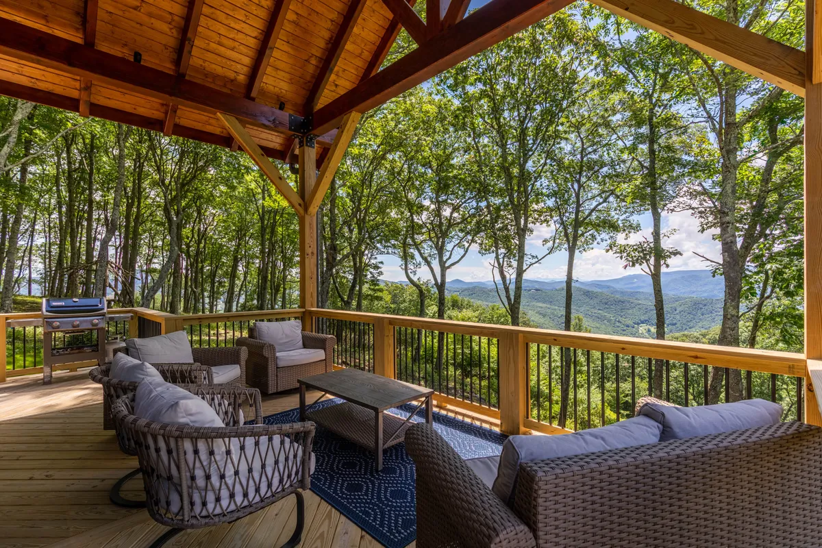 A beautiful outdoor seating area set on a multi-level wooden deck, framed by sturdy wooden beams and a built-in grill. Expansive mountain views stretch out in the background, while comfortable lounge furniture offers a restful retreat for dining or relaxation. The soft golden light of sunset illuminates the deck, highlighting the tranquil atmosphere and connection to nature.