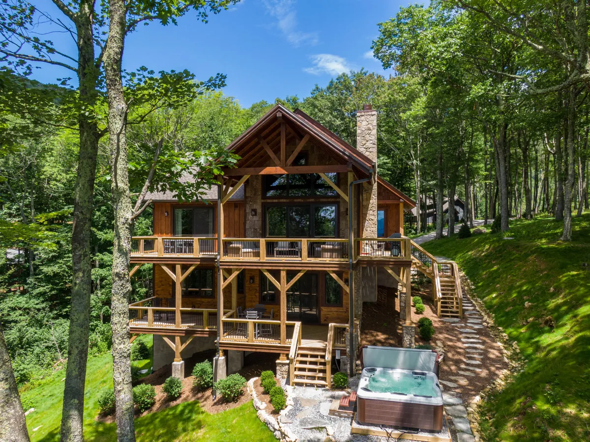 The image showcases the exterior of a custom-built mountain home nestled among towering trees, featuring a spacious wooden deck with a bubbling hot tub, large windows offering panoramic views, and a striking stone chimney. The peaceful scene suggests opportunities for relaxing soaks, outdoor dining, and gathering with loved ones while taking in the surrounding Blue Ridge Mountains.