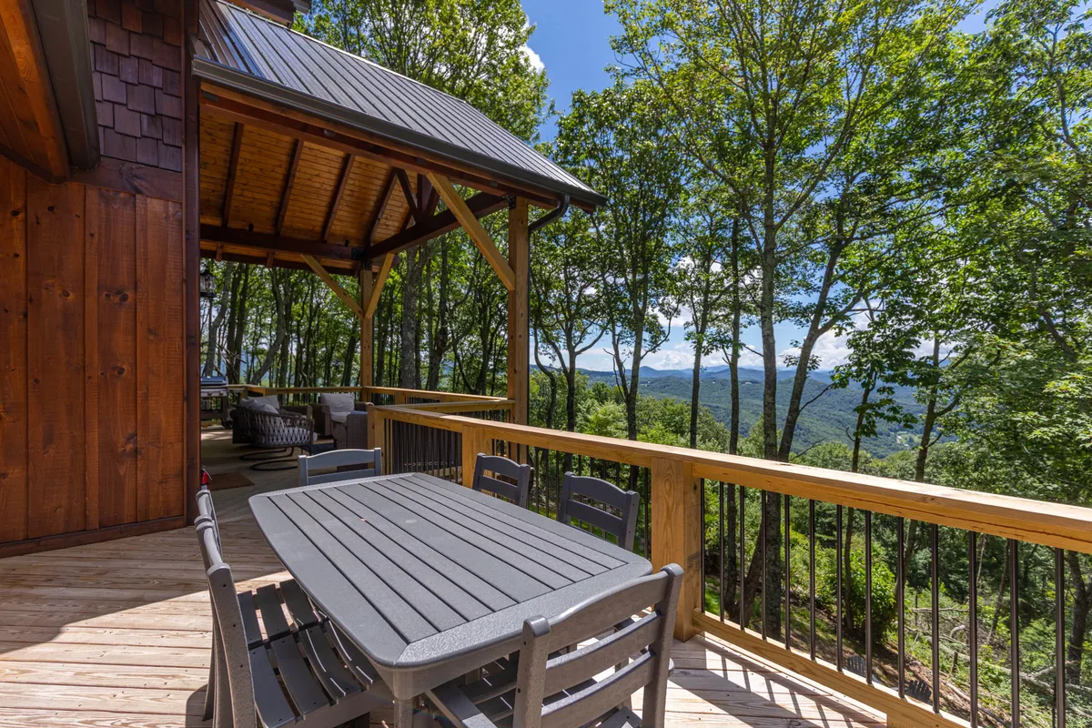 A welcoming wooden deck with a dining table set for outdoor meals, surrounded by lush trees and offering expansive mountain views. The space feels open to nature, perfect for relaxing or letting dogs lounge while their owners enjoy a meal.