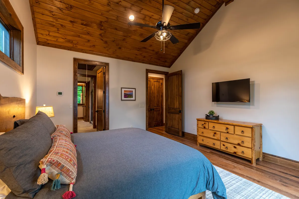 A spacious bedroom featuring a large bed, wooden ceiling, dresser, and wall-mounted TV. The room is bathed in soft, natural light, creating a calm and comfortable space ideal for relaxation and downtime.
