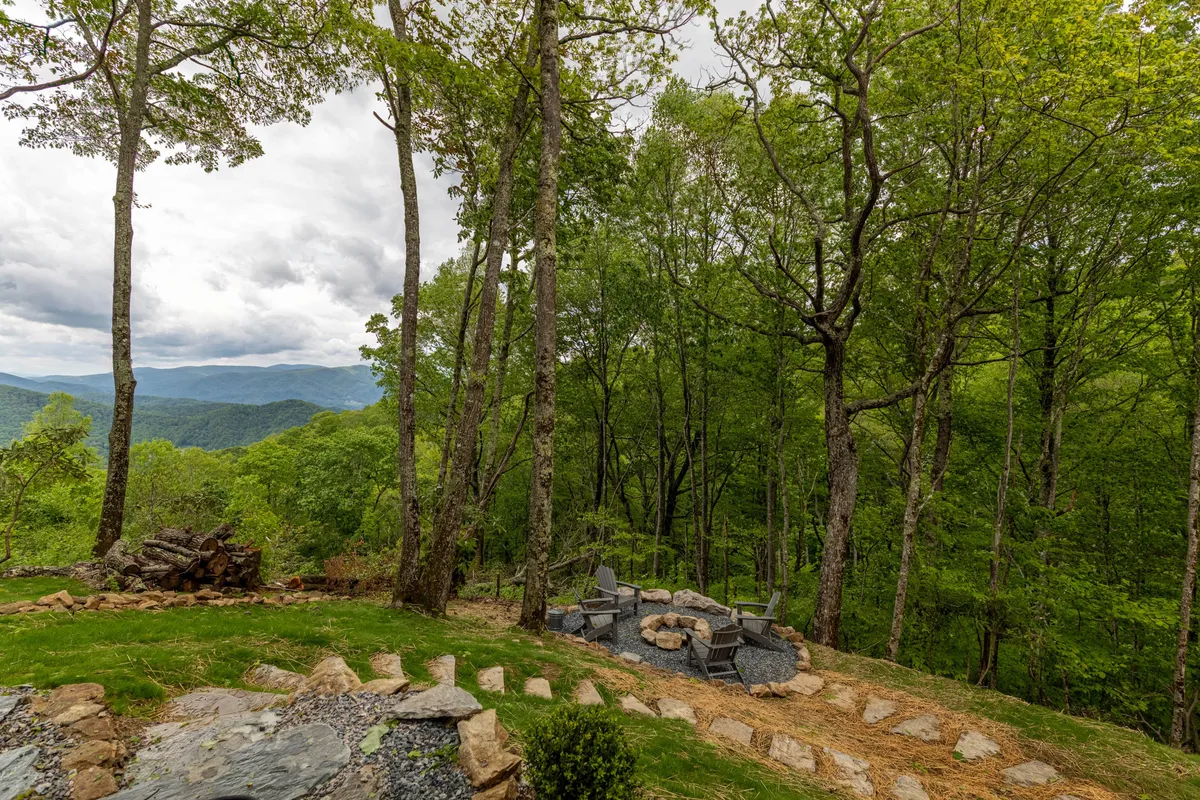 A cozy fire pit area with Adirondack seating is tucked amidst mature trees, with a winding stone pathway leading through the natural landscape. In the distance, mountain peaks rise and the setting sun casts a warm glow, hinting at memorable evenings outdoors.