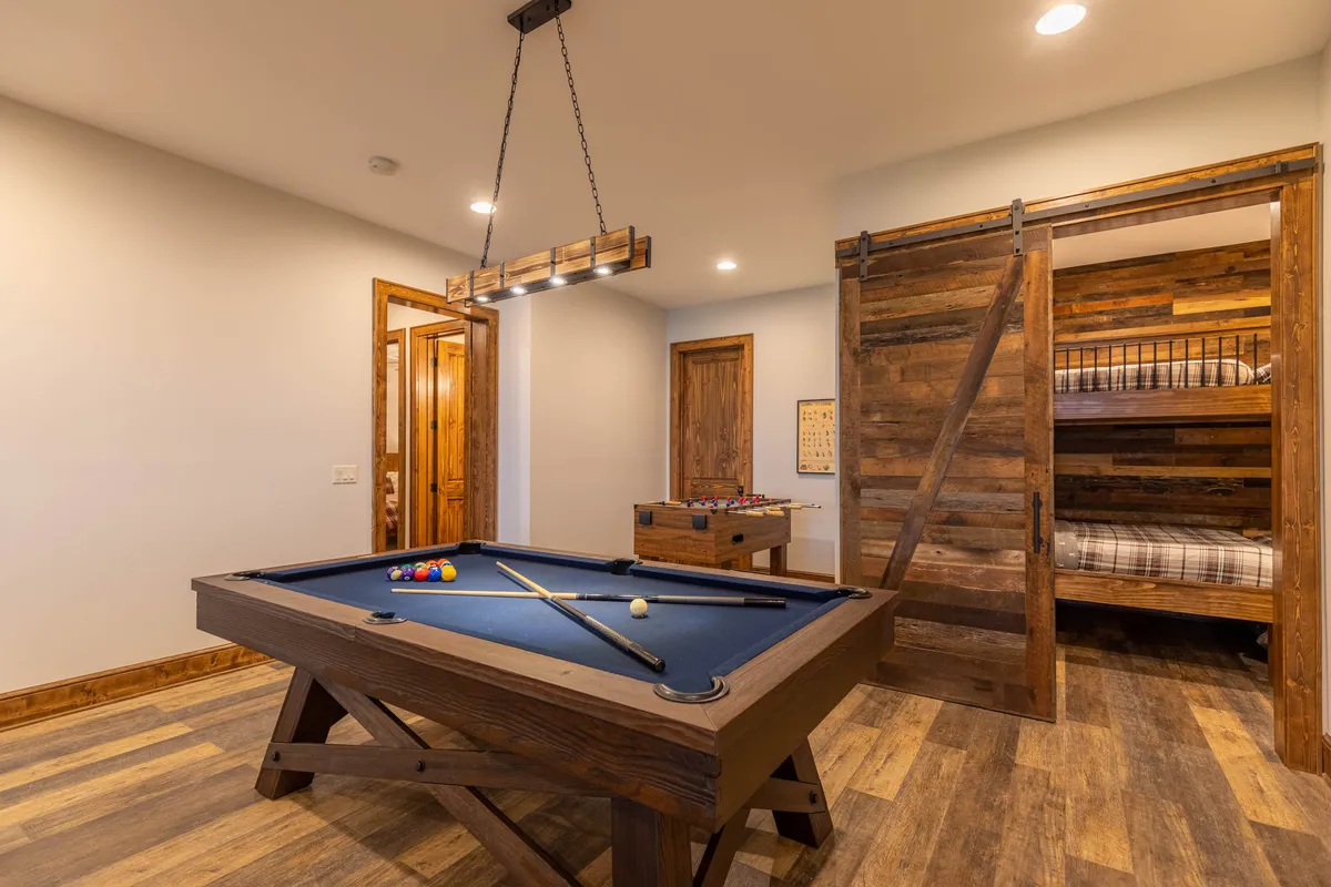 A lively game room featuring a pool table and foosball table situated beside a set of cozy bunk beds with a wooden sliding door in the background. The open arrangement encourages both active play and relaxing downtime, creating an inviting multi-purpose entertainment hub for guests of all ages.