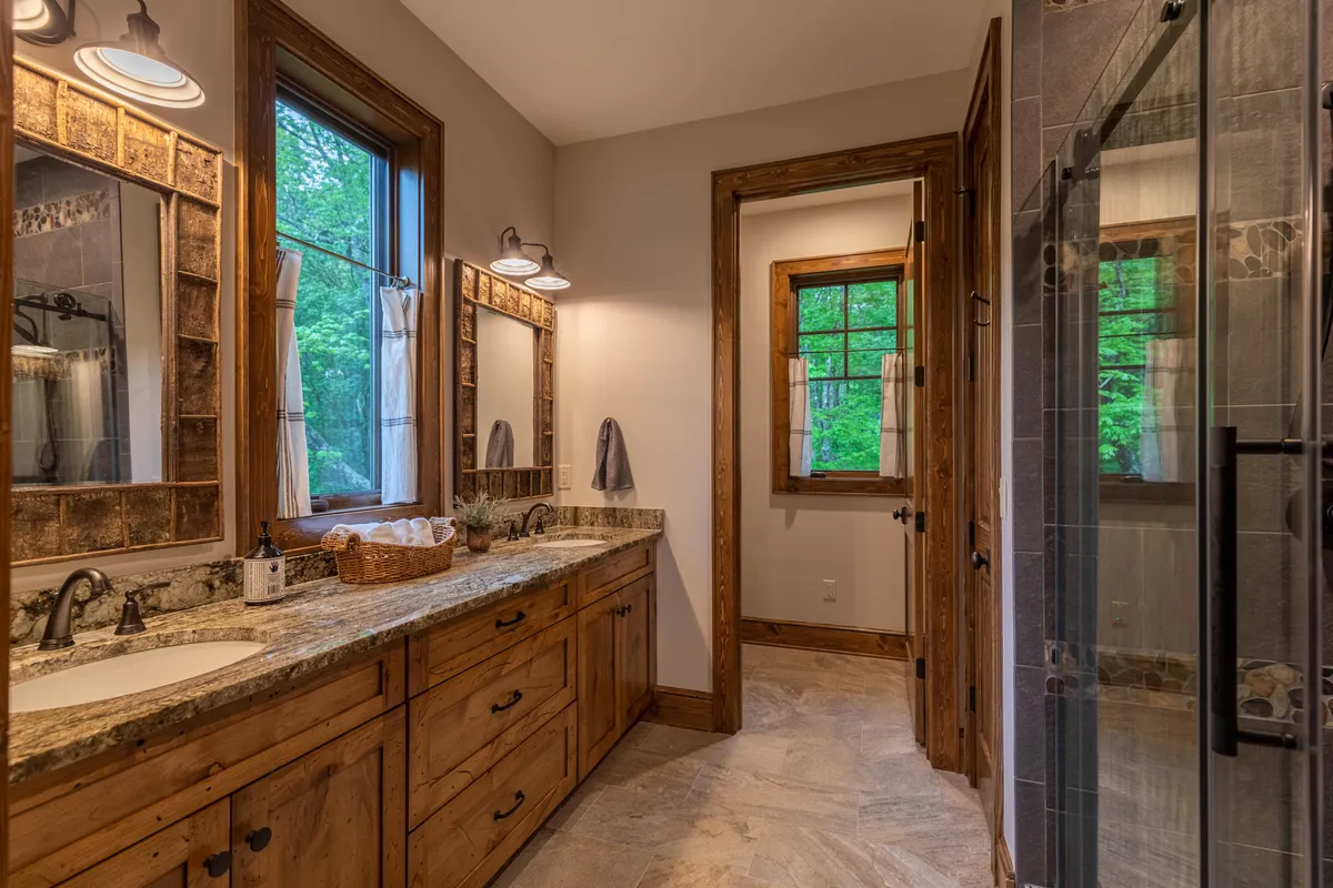 A contemporary ensuite bathroom with a double sink vanity, large mirrors, a shower enclosure, and windows offering natural views. The mix of custom tile, granite finishes, and abundant light creates a spa-like ambiance.