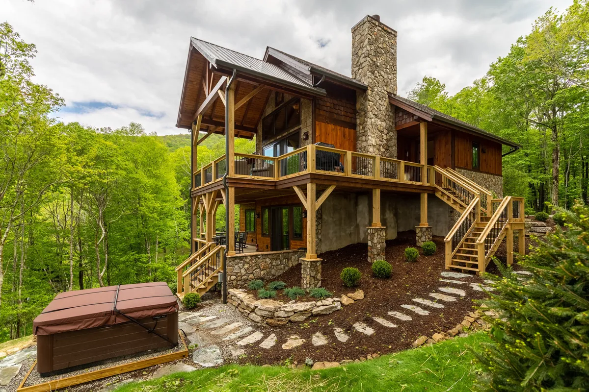 A spacious wooden deck wraps around the home, featuring a bubbling hot tub and a stone chimney. Lush greenery surrounds the space, creating a serene and welcoming environment for group gatherings and outdoor relaxation.